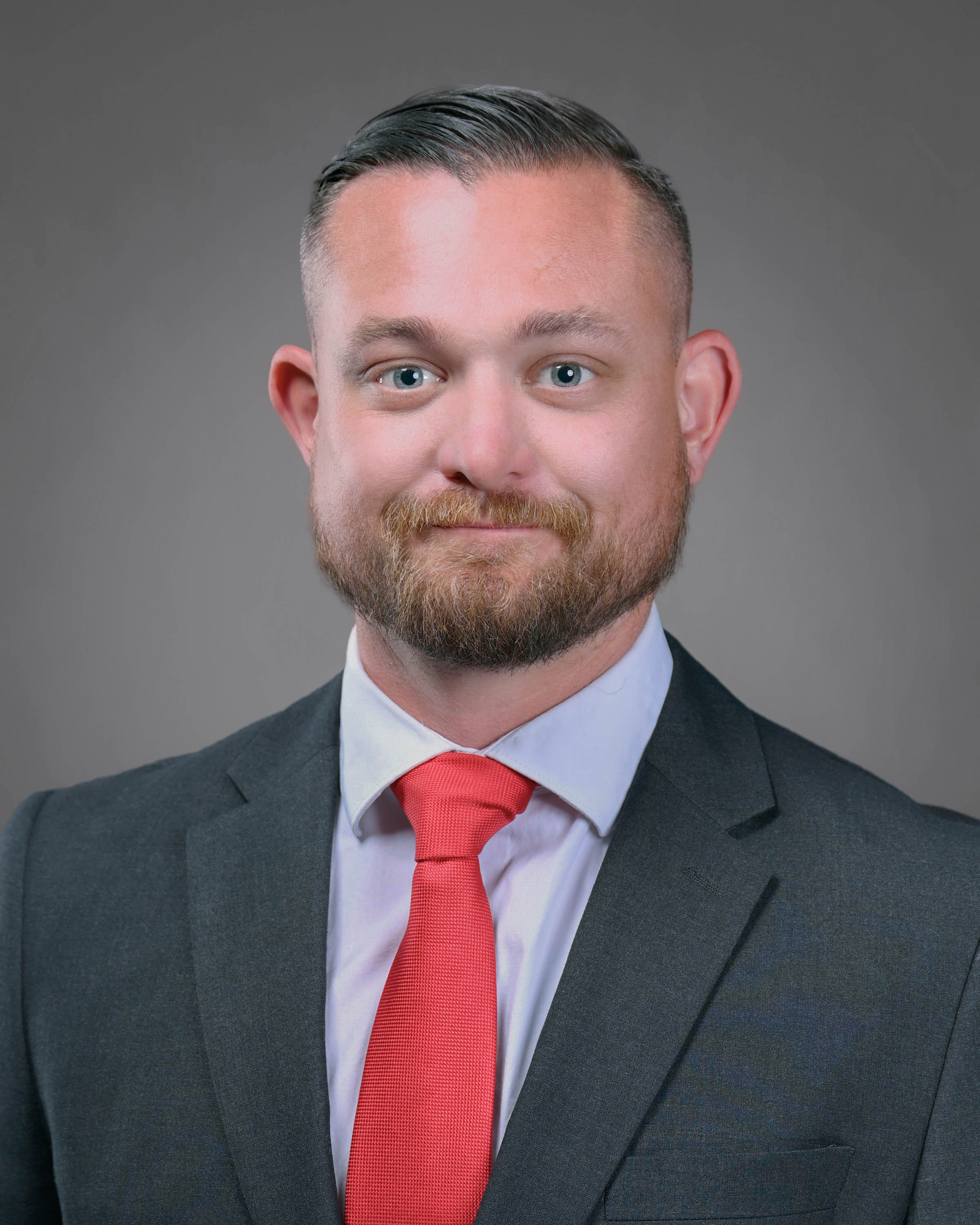 Man with short hair and a beard smiling with a dark suit jacket and red tie